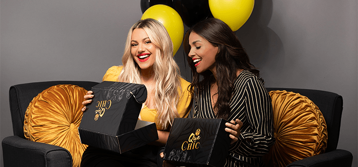 Dos mujeres sonrientes abriendo dos cajas Qué Chíc de color negro y dorado
